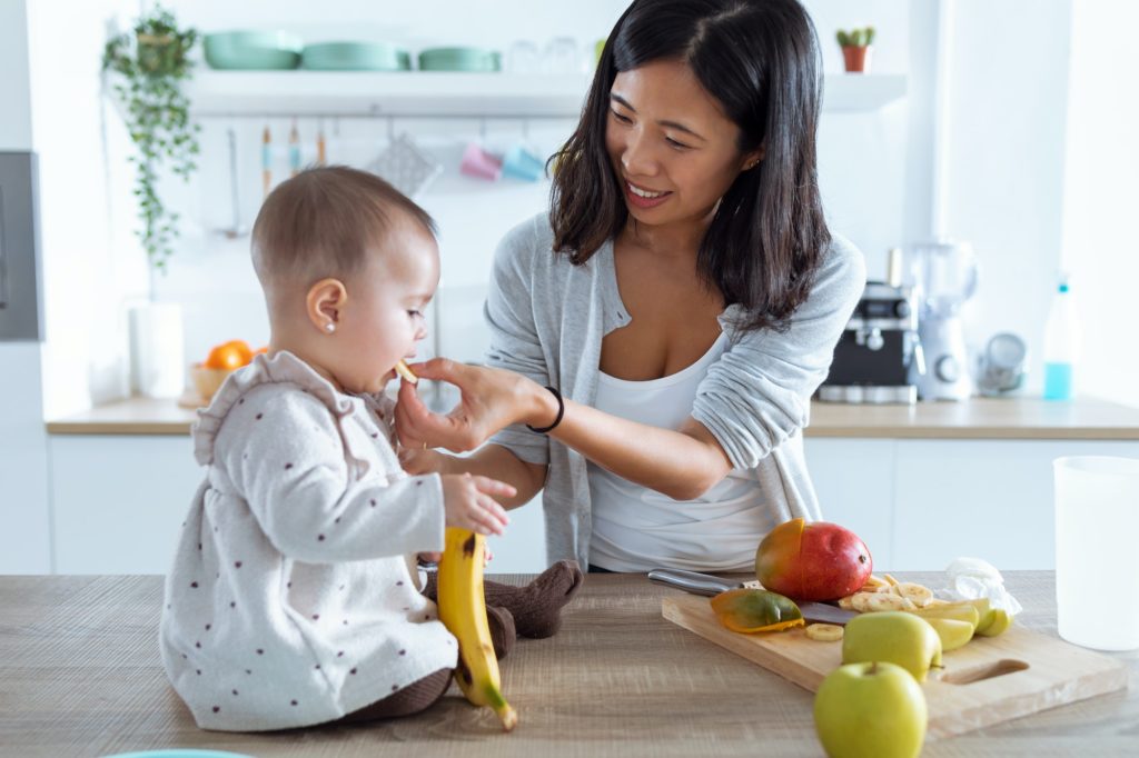 Happy young mother feeding her cute baby girl with a banana in the kitchen at home.